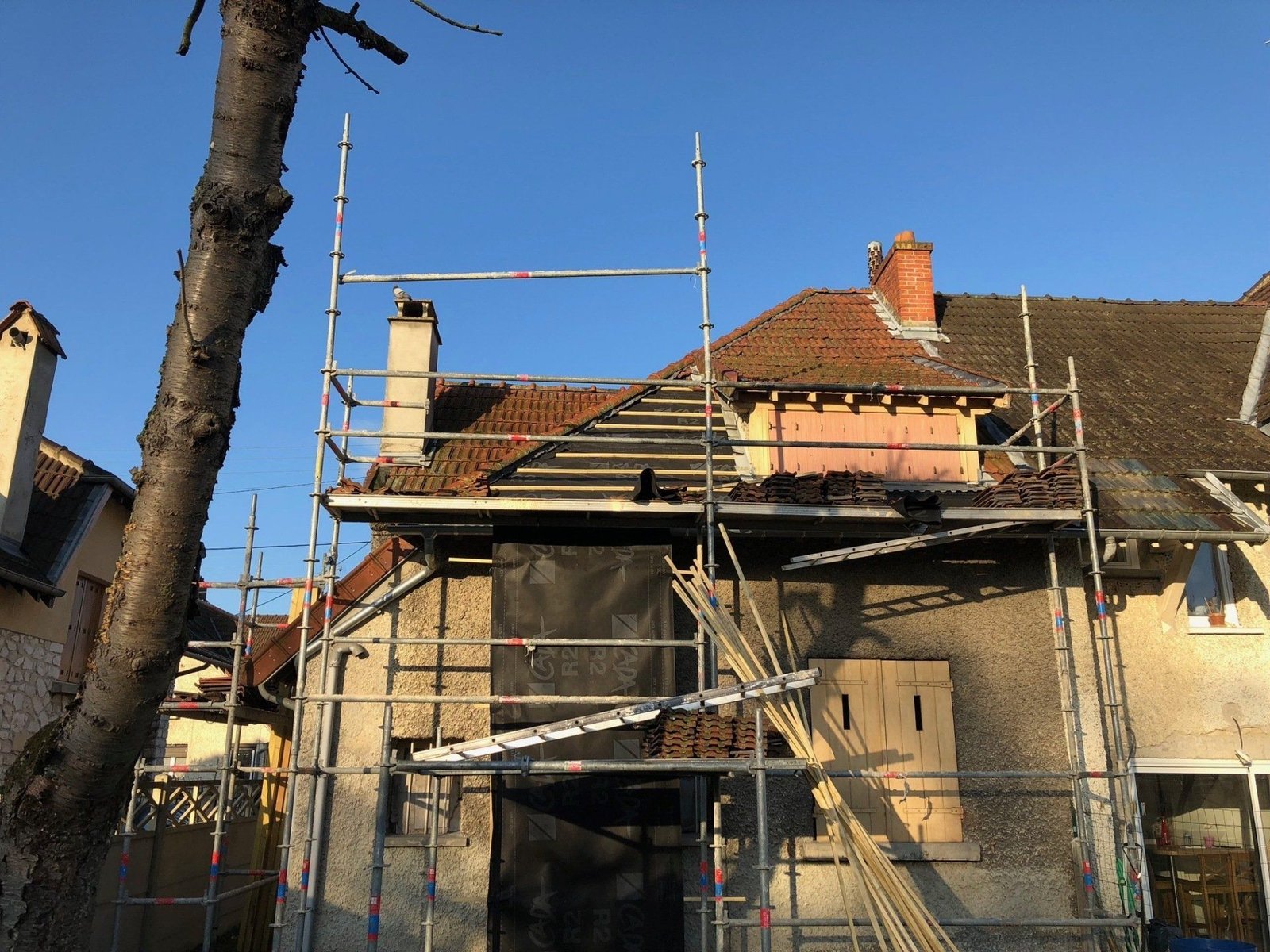 Une maison est en cours de rénovation avec des échafaudages et un arbre au premier plan.